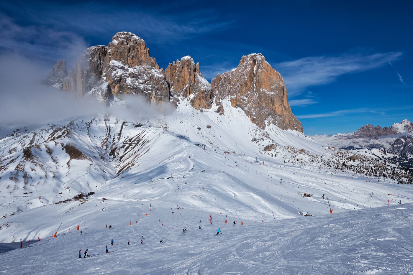 Skiers on a wide piste below jagged rock peaks