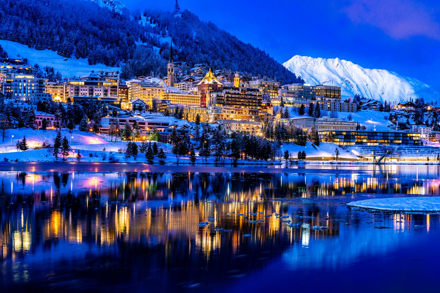 A lakeside town lit up at dusk below snowy mountains